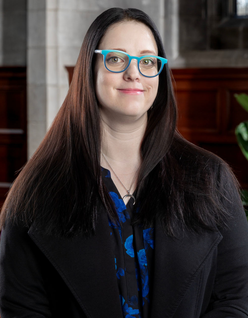 A woman with long dark hair, wearing blue glasses and a black coat, poses for a portrait against a backdrop of wooden panels and architectural details.