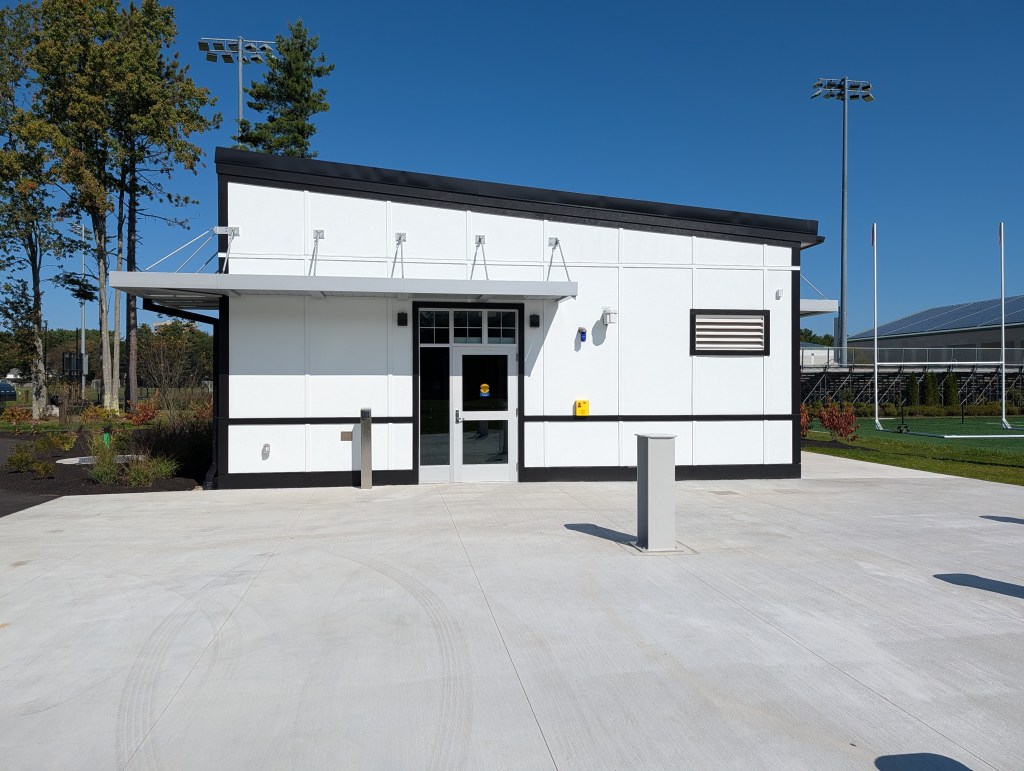 A modern white building with a black trim, featuring a glass entrance, surrounded by greenery and sports field equipment.