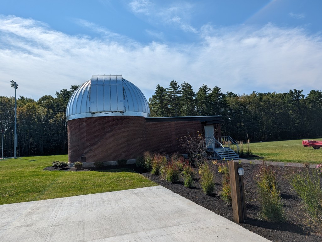 A brick observatory building with a silver dome, located in a green field with trees in the background and a clear blue sky.